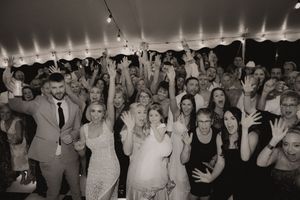 Black and white photo of a large wedding reception group celebrating under a tent with hands raised joyfully.