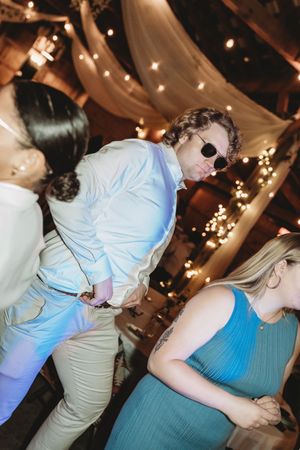 Guests dancing at a wedding reception in a warmly lit barn decorated with string lights and draped fabric.