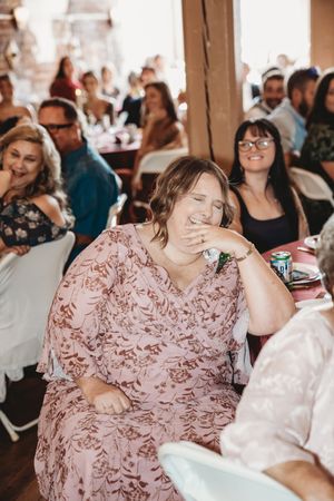 Woman in floral pink dress laughing at a wedding reception with guests seated in background.