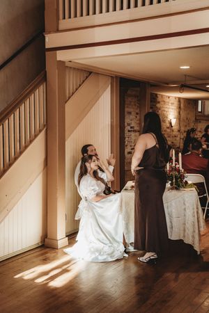 Bride in white gown seated near staircase chatting with bridesmaid in brown dress at rustic wedding venue.