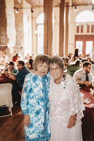 Two elderly women smiling together at a wedding reception in a rustic venue with guests seated in the background.
