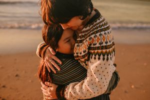Couple embracing on a beach at sunset wearing cozy sweaters against an orange sky and sandy backdrop.