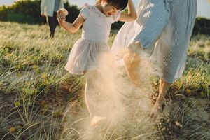 A young girl in a pink tutu playfully kicks up sand while walking barefoot through the grassy dunes at Kurnell Beach.
