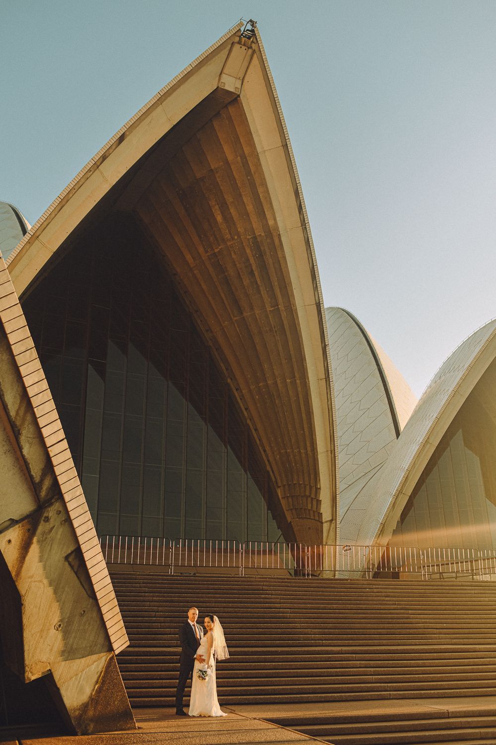 A newlywed couple posing on the steps of the Sydney Opera House, with the iconic architectural sails towering above them
