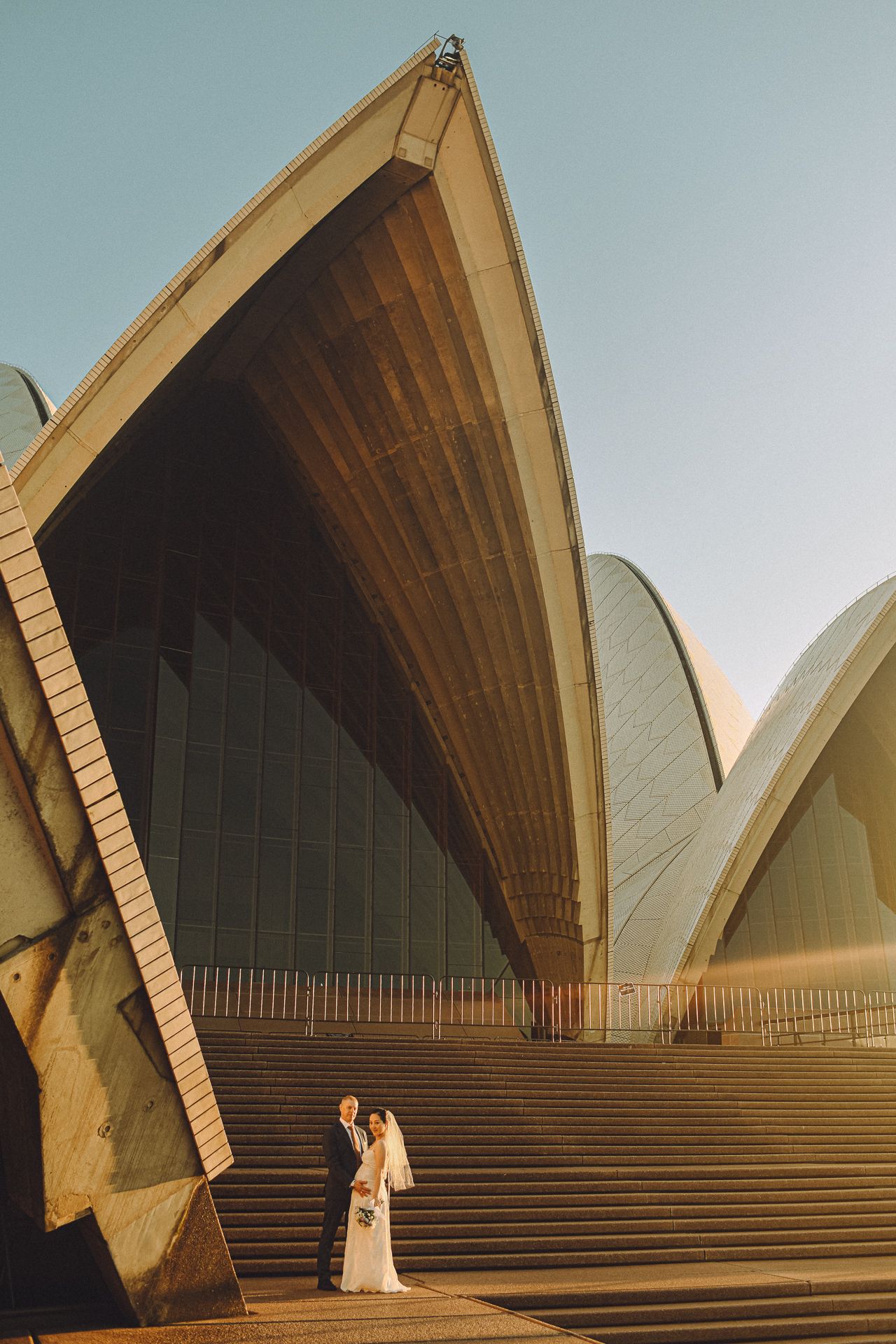 A newlywed couple posing on the steps of the Sydney Opera House, with the iconic architectural sails towering above them