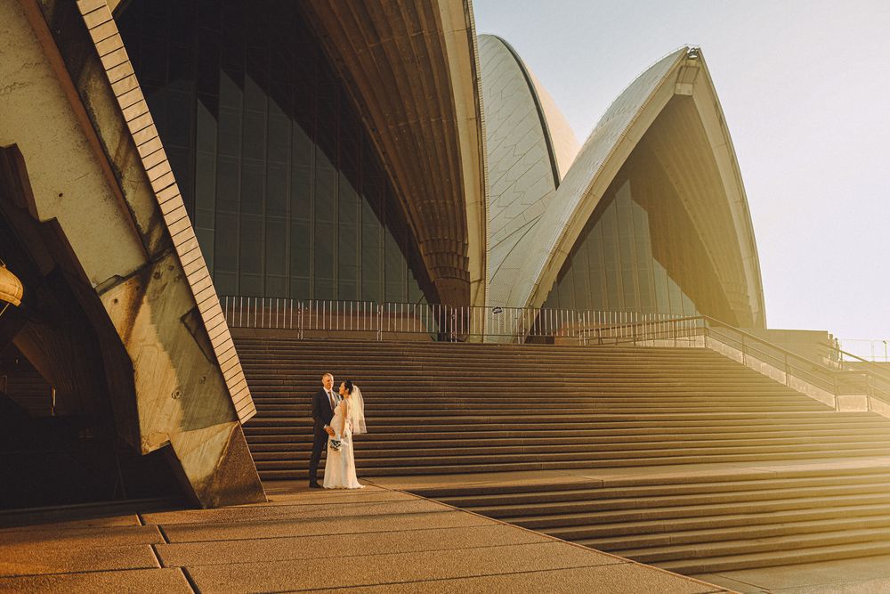 A bride and groom standing together on the steps of the Sydney Opera House, bathed in sunrise. The Opera House's