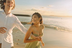 A joyful couple running along the shore at La Perouse Beach during sunset. The woman, wearing a dress, is laughing