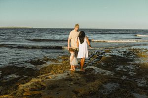 A family of three walking along the rocky shoreline at Cronulla Beach. The parents hold their young daughter's hands