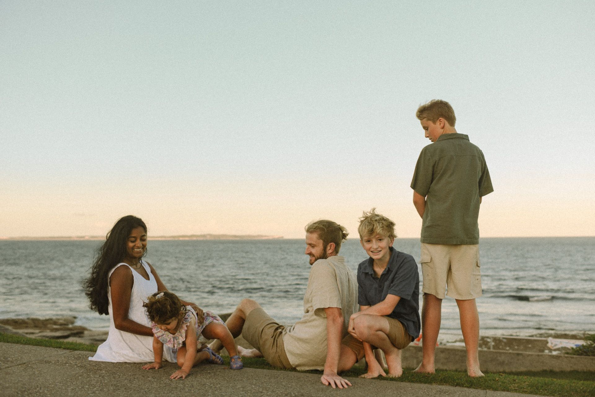 A family sitting together on a grassy cliffside at Cronulla, overlooking the ocean at sunset.