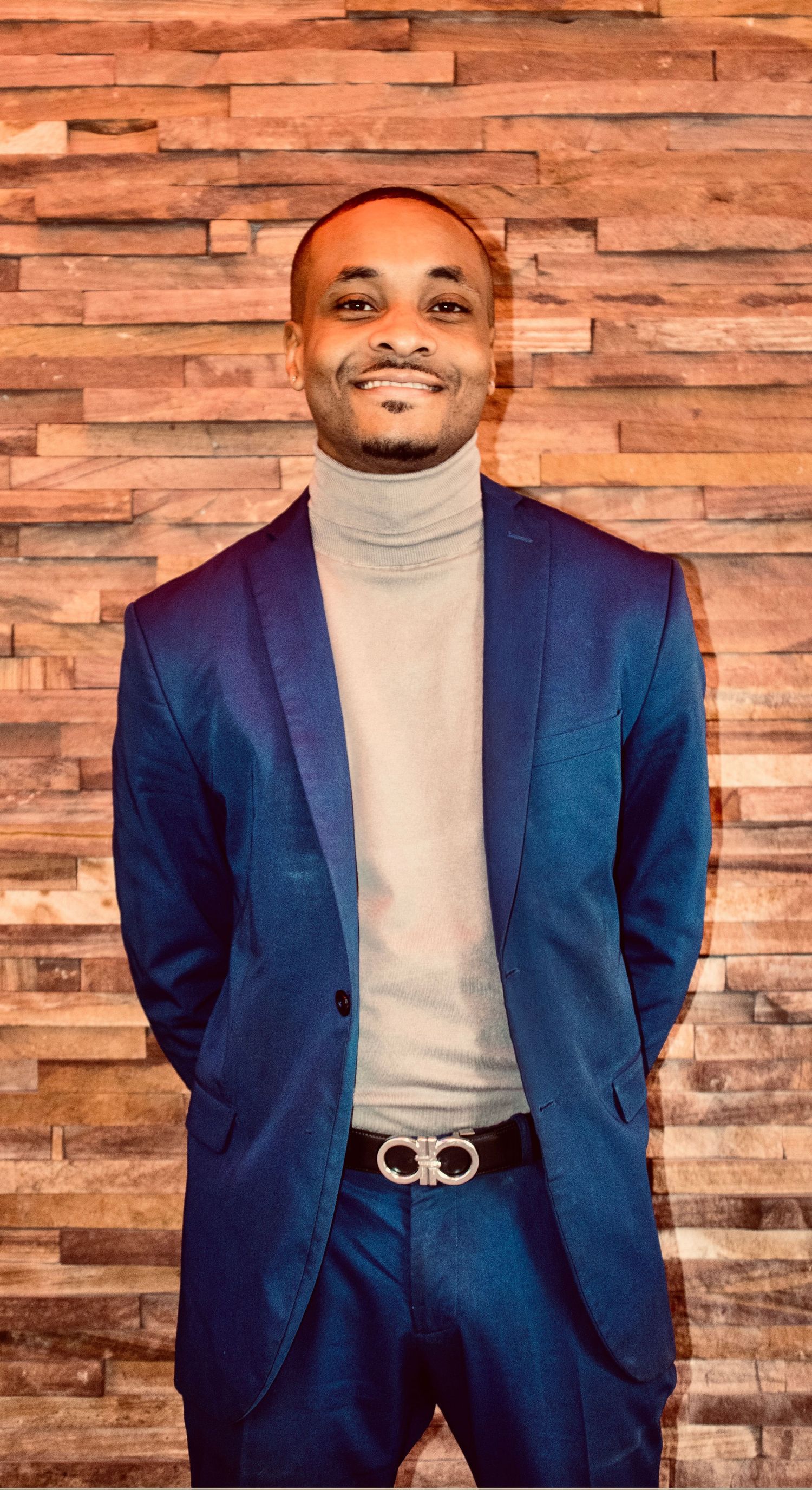 Cheerful man in blue suit and gray turtleneck with silver designer belt smiles against wood wall.