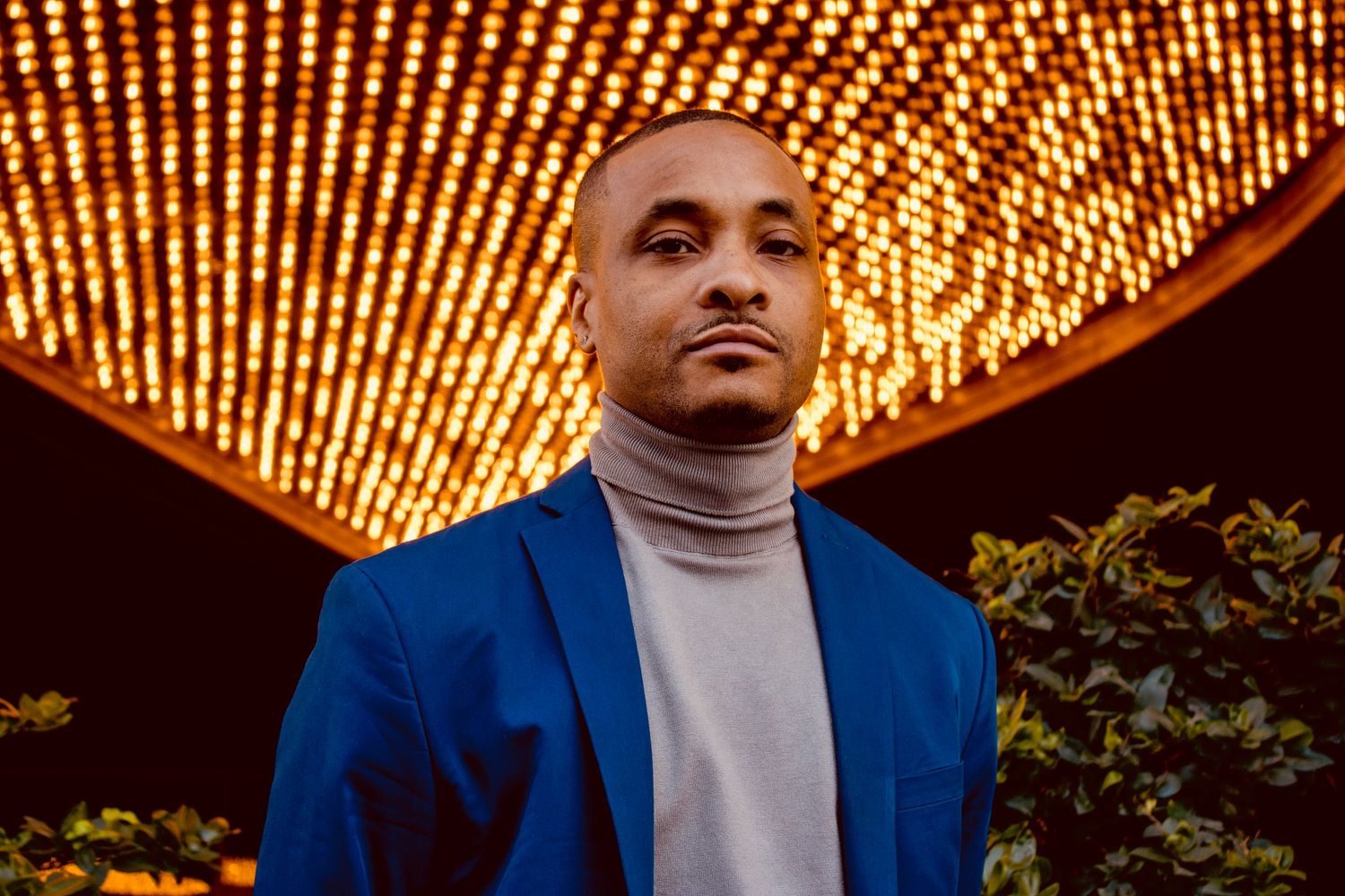 Bald man in blue blazer and gray turtleneck poses under glowing lights.