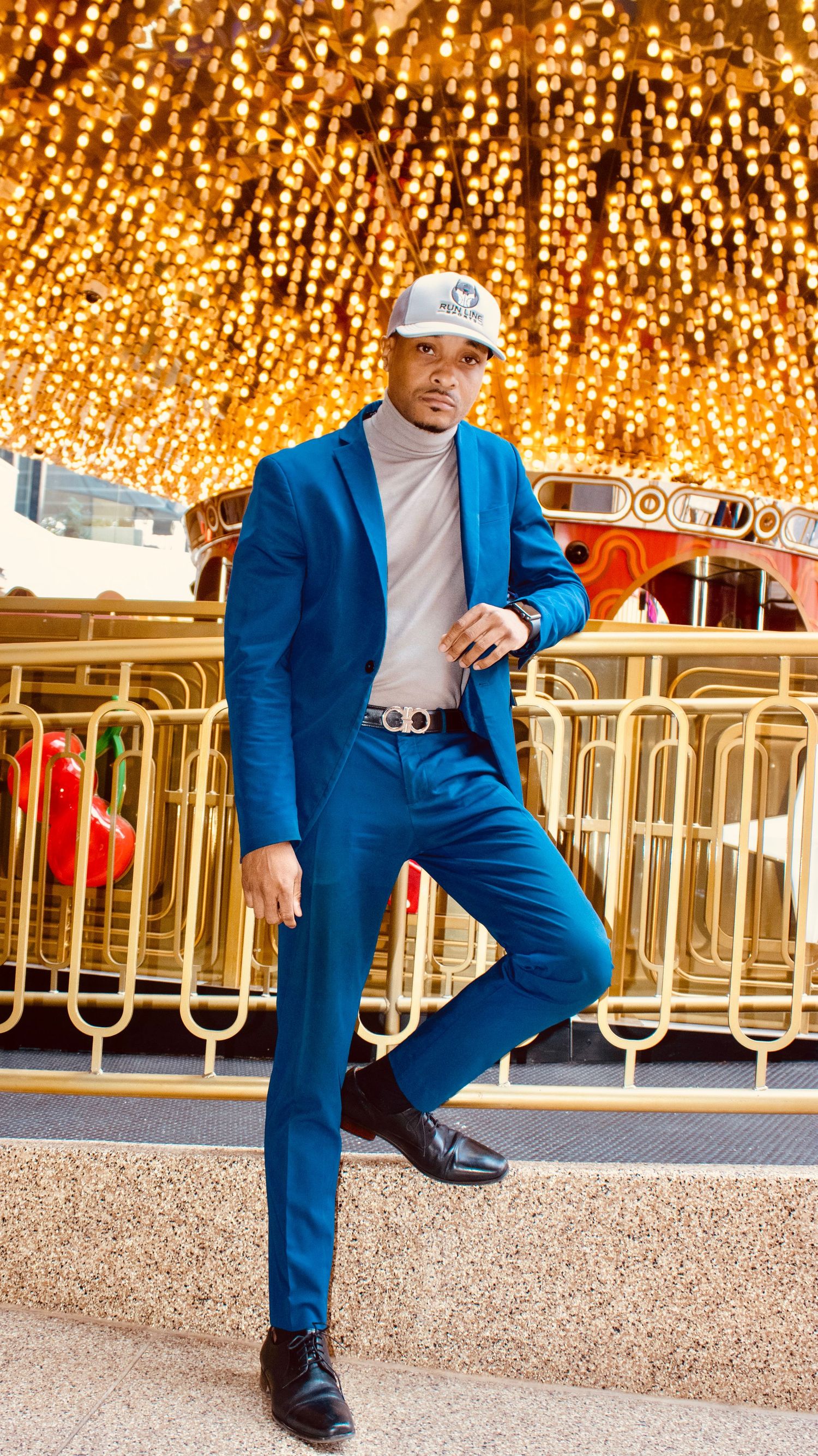 Stylish man in blue suit and white cap poses under golden lights.