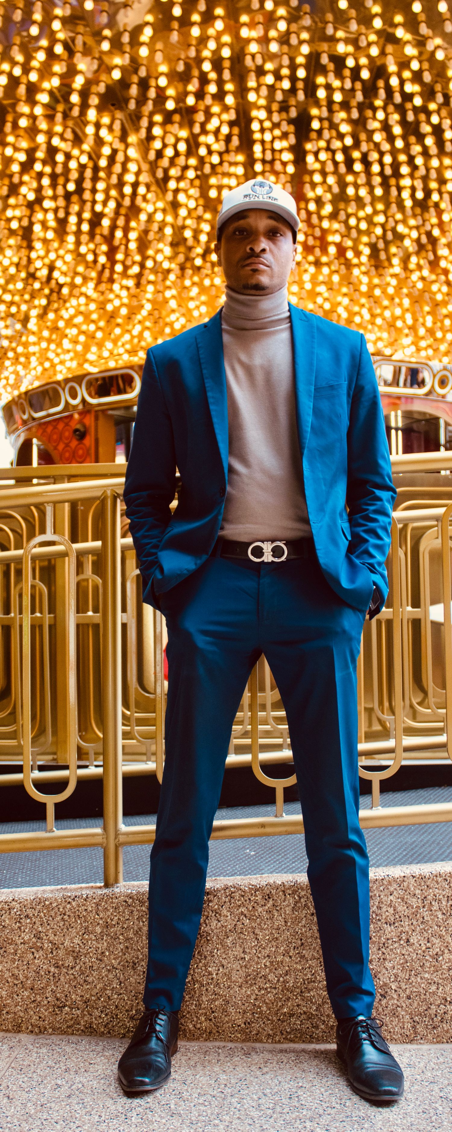 Man in a blue turtleneck suit with designer belt poses confidently under cascading golden lights.