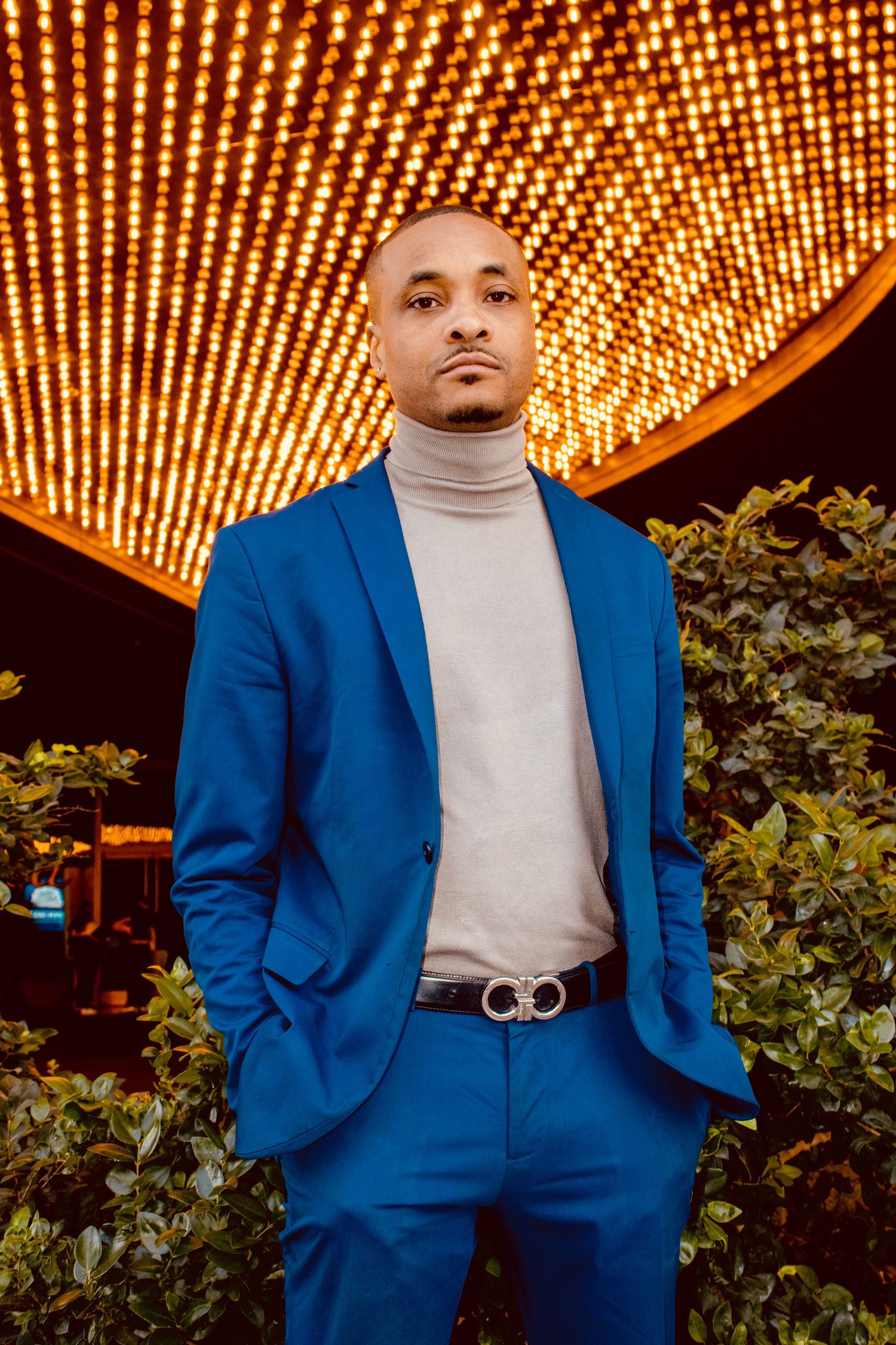 Man in blue suit and gray turtleneck stands before bright marquee lights.