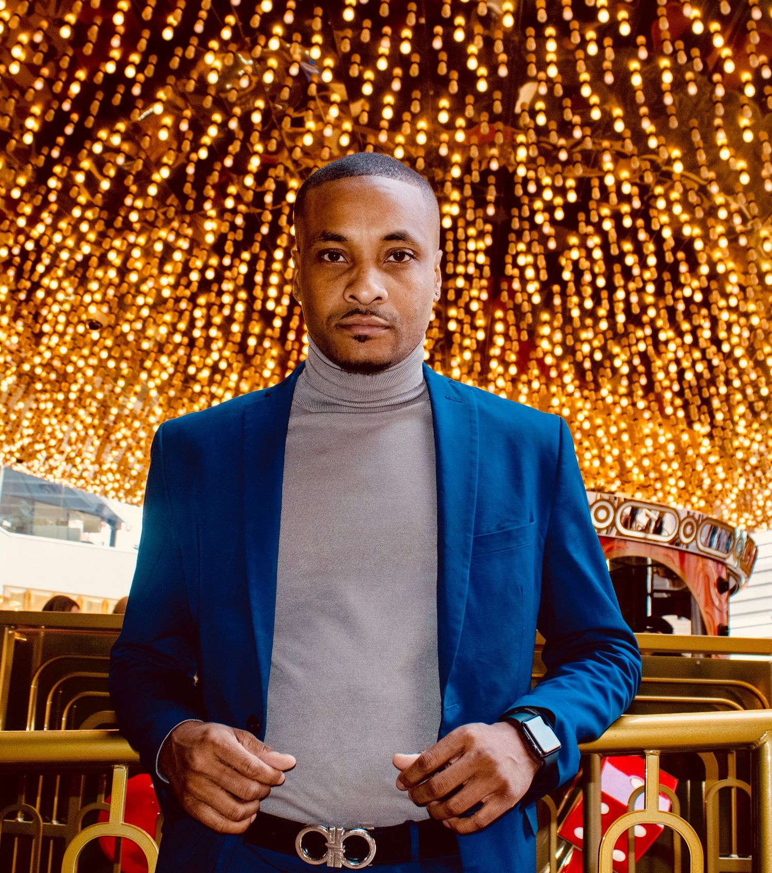 Man in blue blazer and gray turtleneck poses confidently under golden lights.