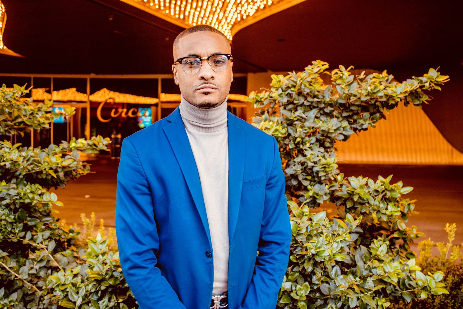 Man in blue suit and turtleneck with glasses stands seriously in front of green bushes outdoors.