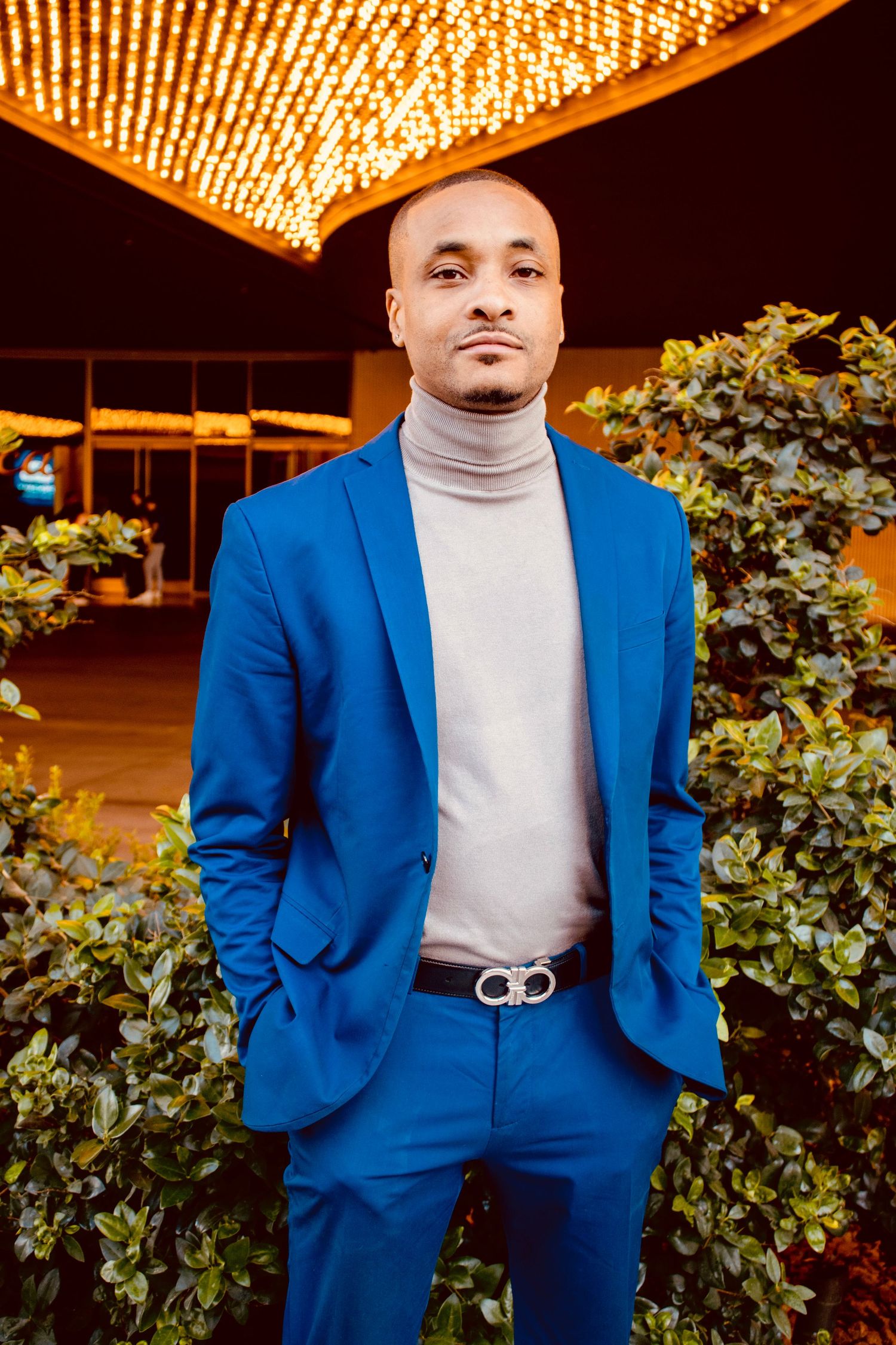 Man in blue suit and turtleneck poses outdoors under golden lights.