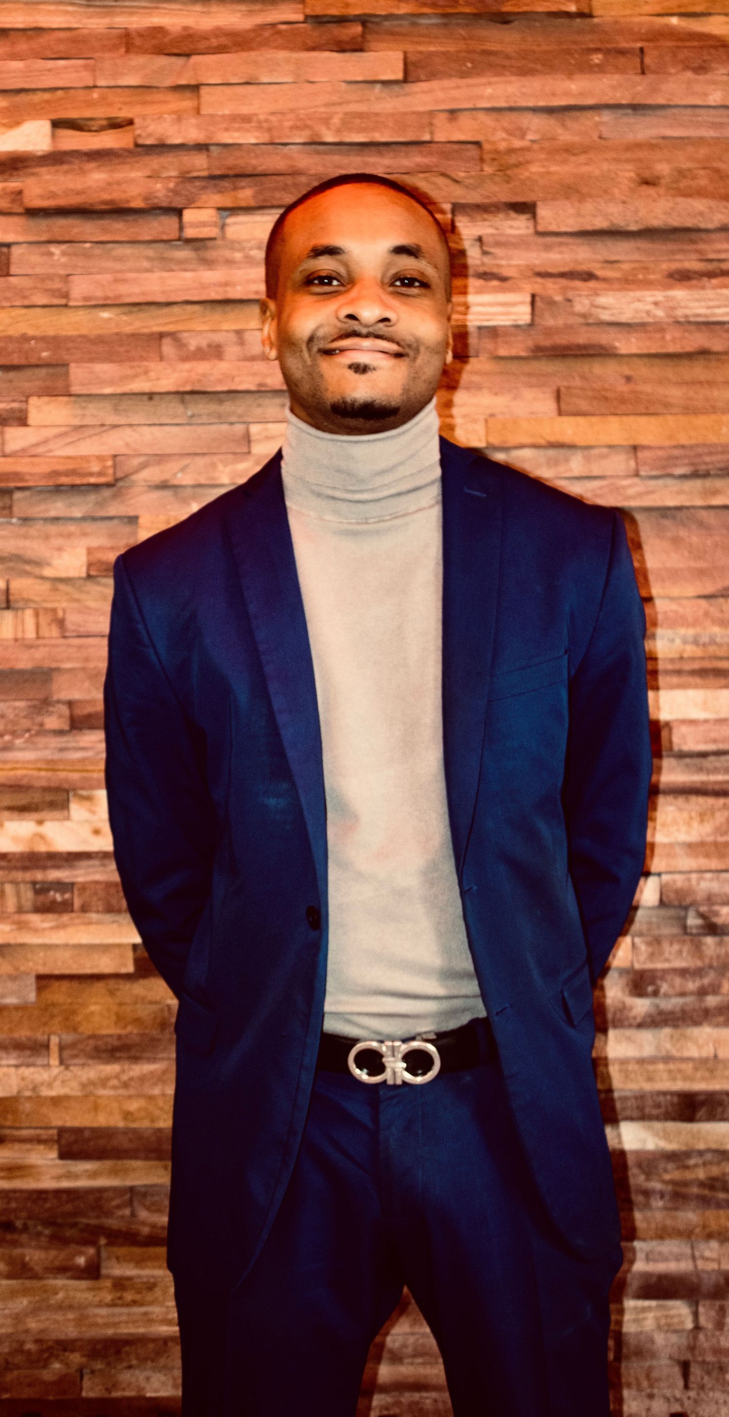 Smiling man in navy blue suit and gray turtleneck with designer belt poses against rustic wood wall.