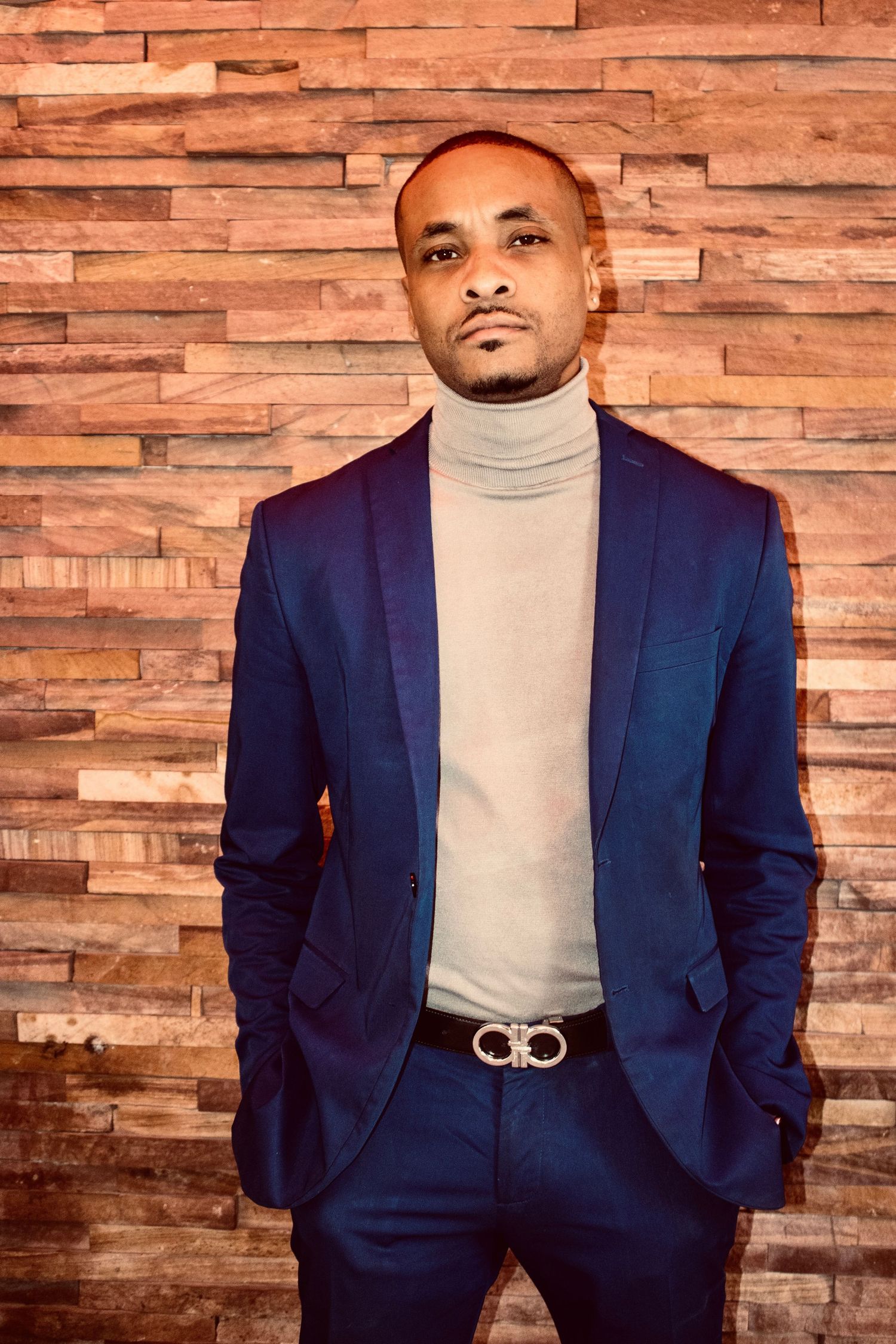 Man in navy blazer and gray turtleneck with designer belt stands against a textured wood panel wall.