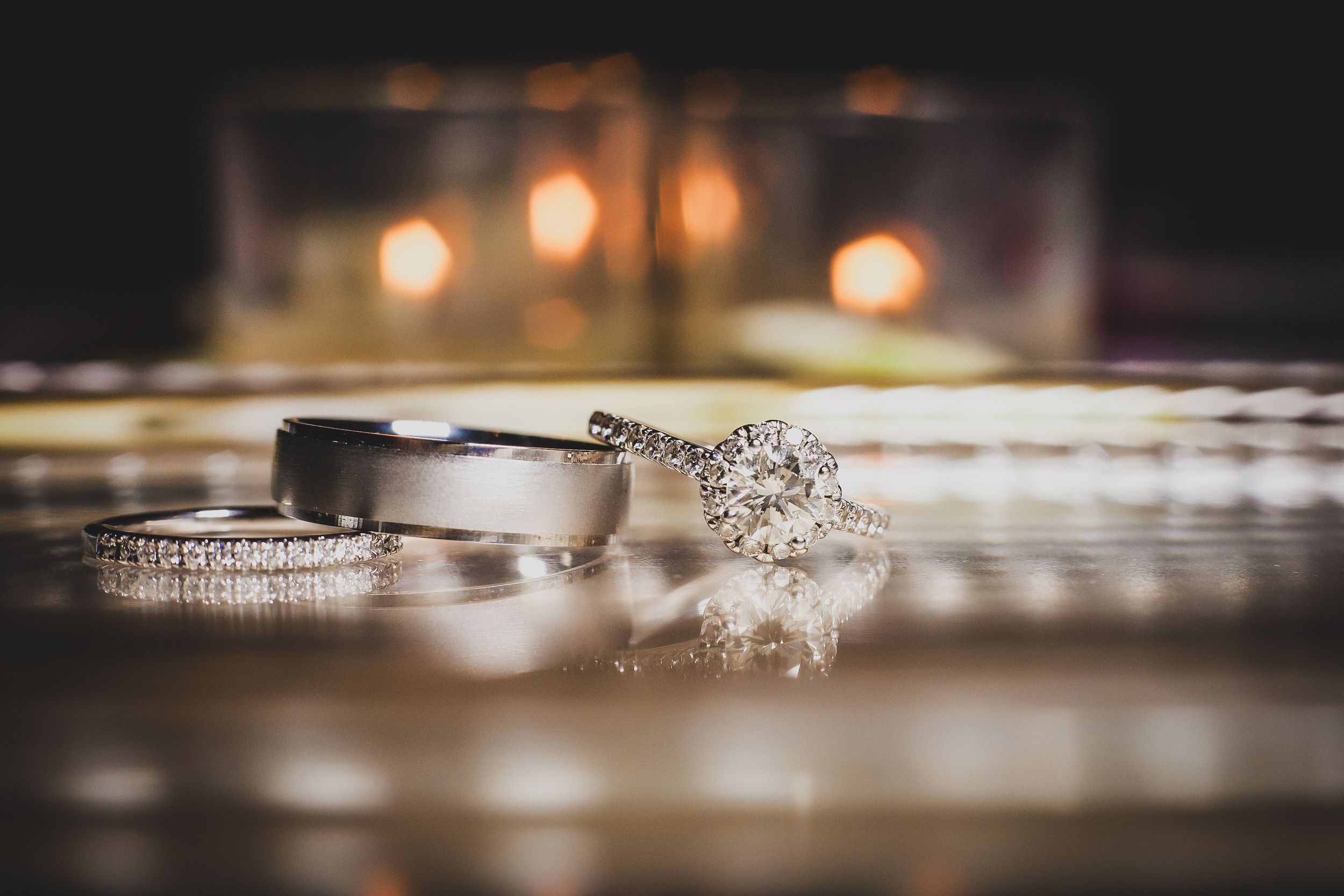 Wedding rings and engagement ring with diamonds sparkle against a reflective surface with soft bokeh lighting in background.
