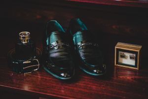 Black leather dress shoes with cologne and cufflinks displayed on dark wooden surface in moody lighting.