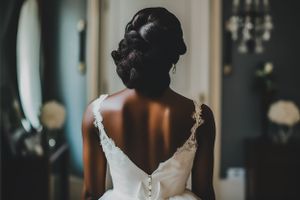 A backlit bride in a white wedding dress with delicate straps stands in a moody, dimly lit room.