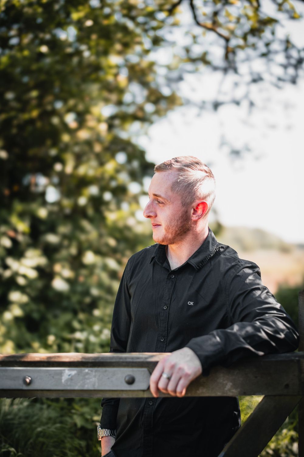 Person in black shirt standing by wooden fence in a natural outdoor setting with sunlit foliage in background.