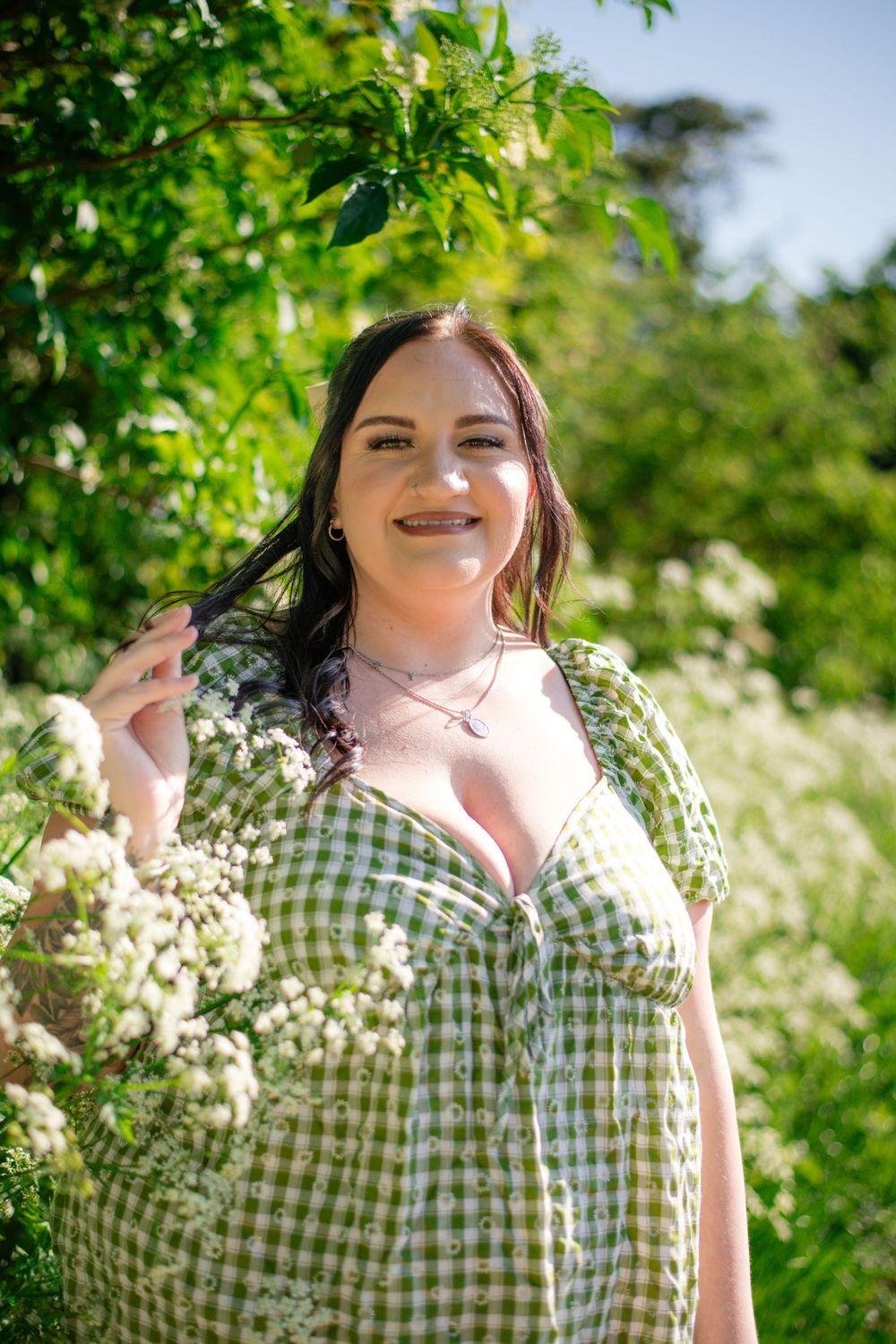 Smiling woman in green gingham dress standing among flowering bushes on sunny day.