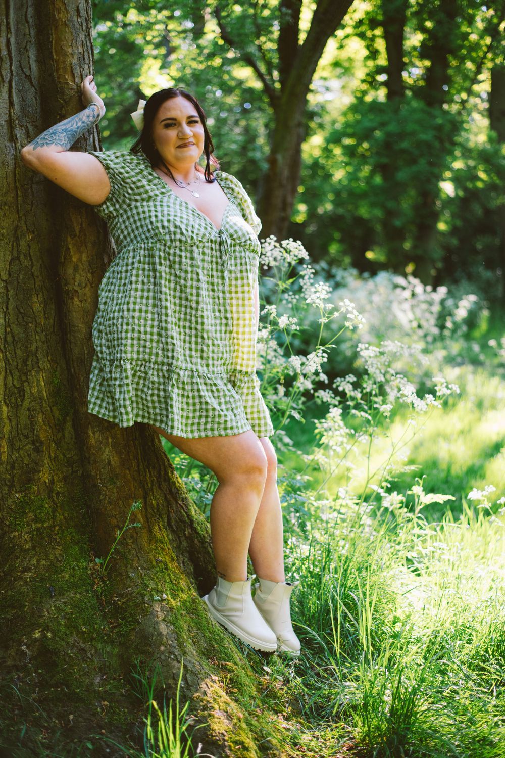 Woman in gingham dress leaning against tree trunk in sunny woodland setting.
