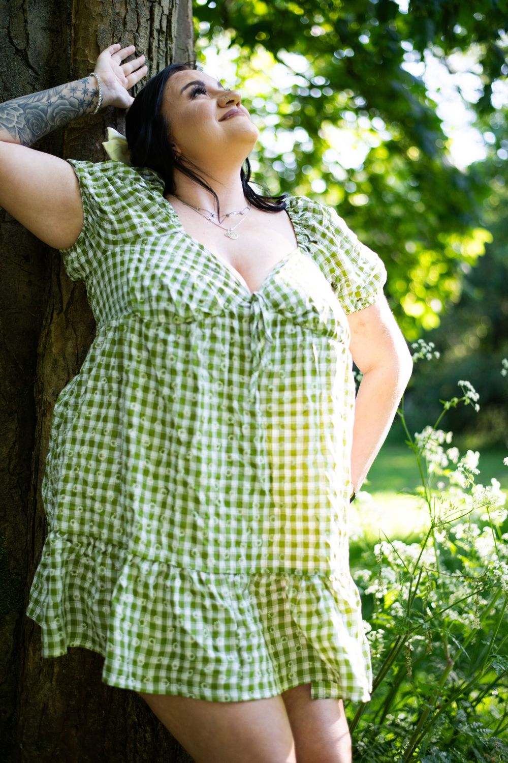 A green gingham dress-clad figure leans against a tree in dappled sunlight outdoors.