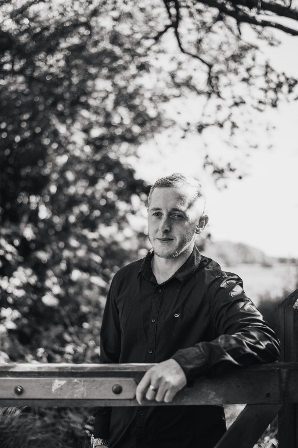 Black and white portrait of person leaning against rustic wooden fence with trees in background.