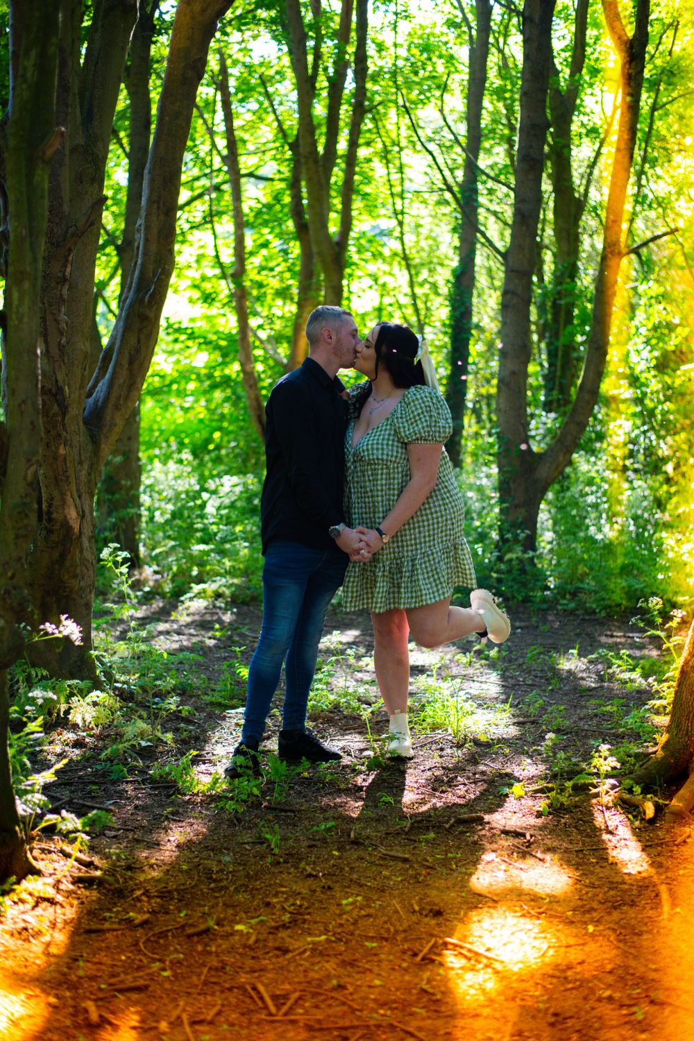 A couple shares an intimate moment on a sunlit forest path wearing coordinated outfits.