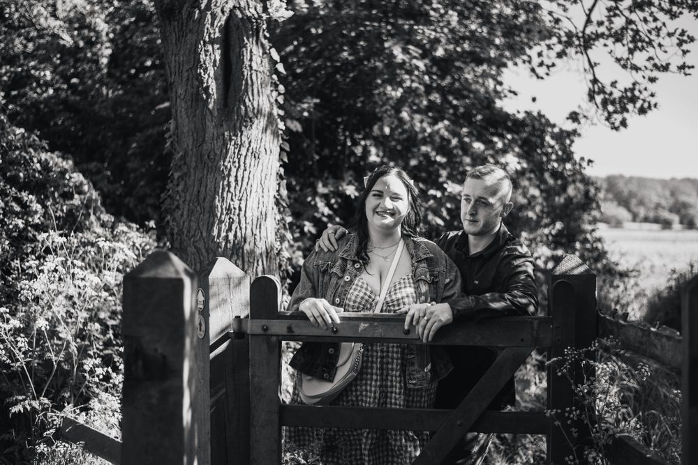 Person in plaid shirt standing at wooden gate in black and white nature setting.