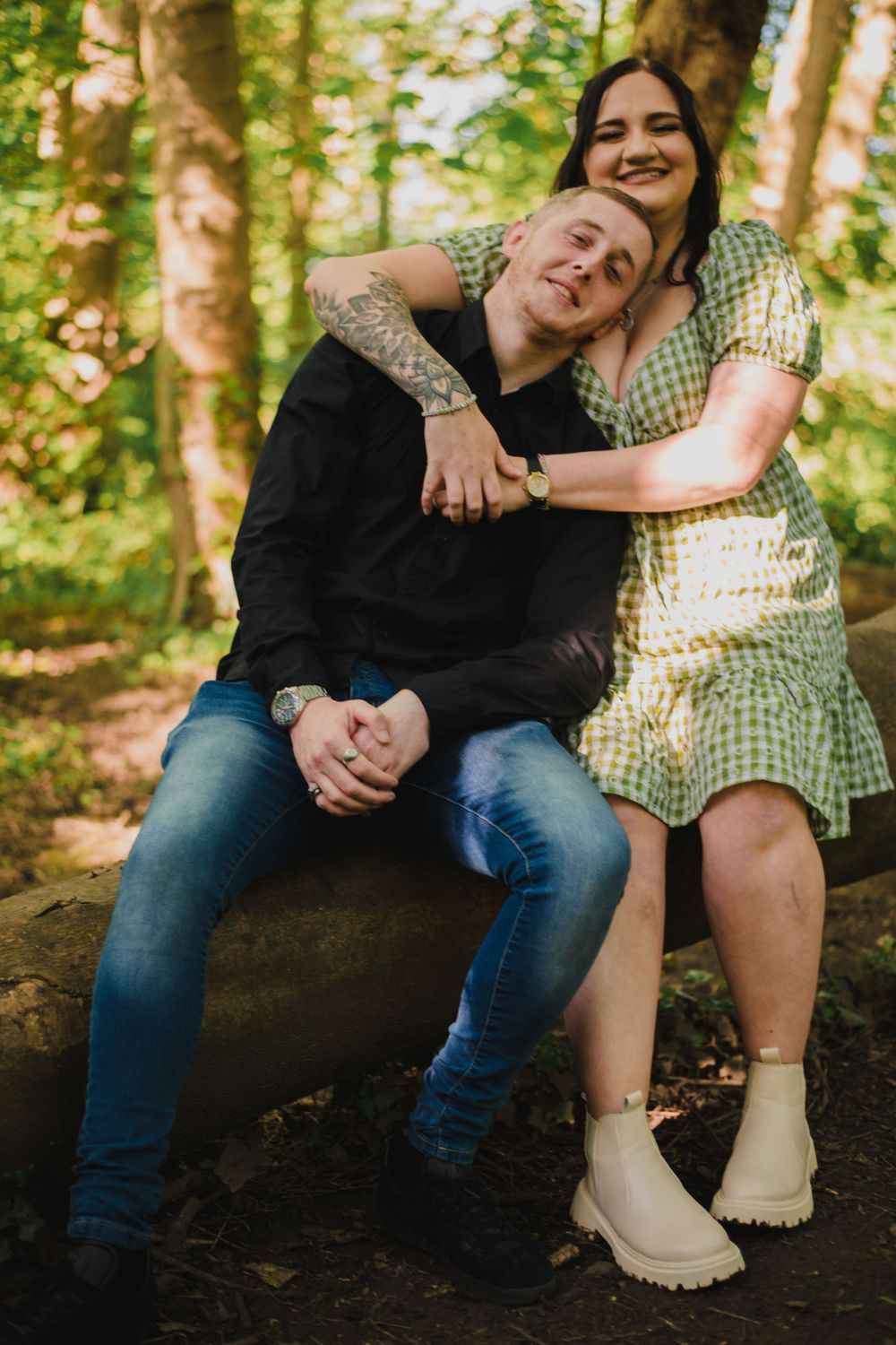 A couple embraces on a log in a sun-dappled forest setting.