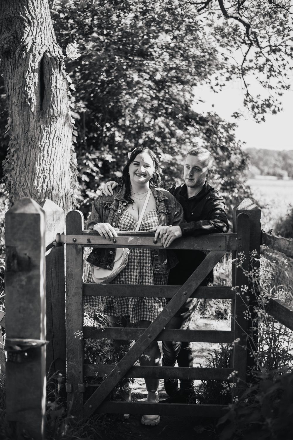 Two people standing at rustic wooden gate in black and white photograph with trees in background.