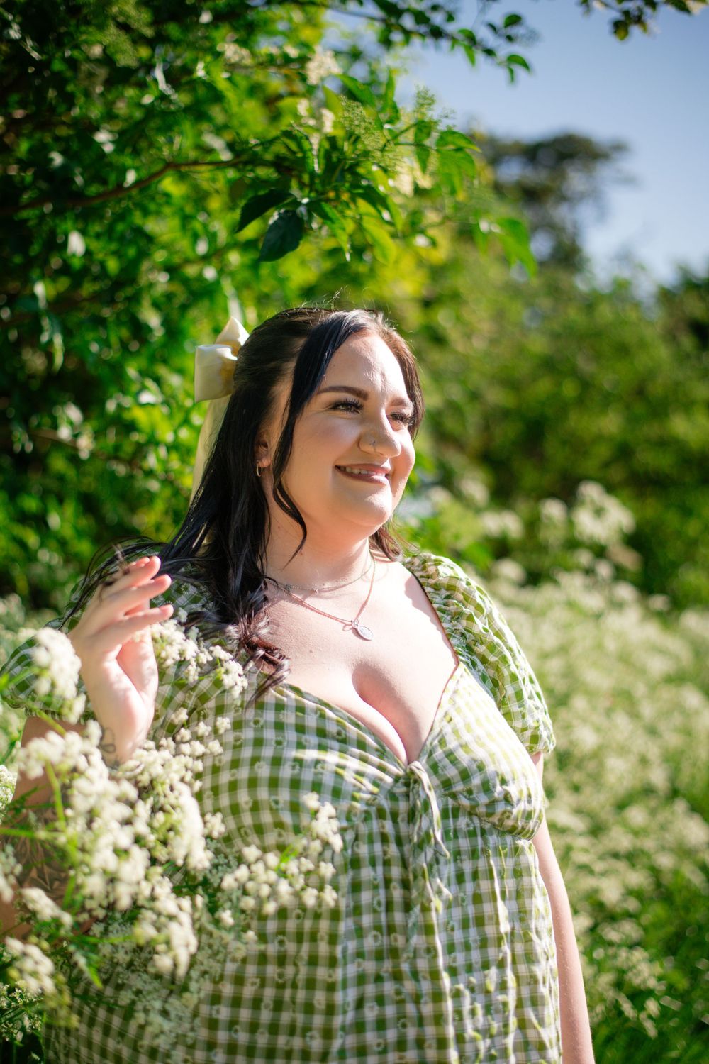 Woman in green gingham dress poses among blooming flowers and greenery on sunny day.