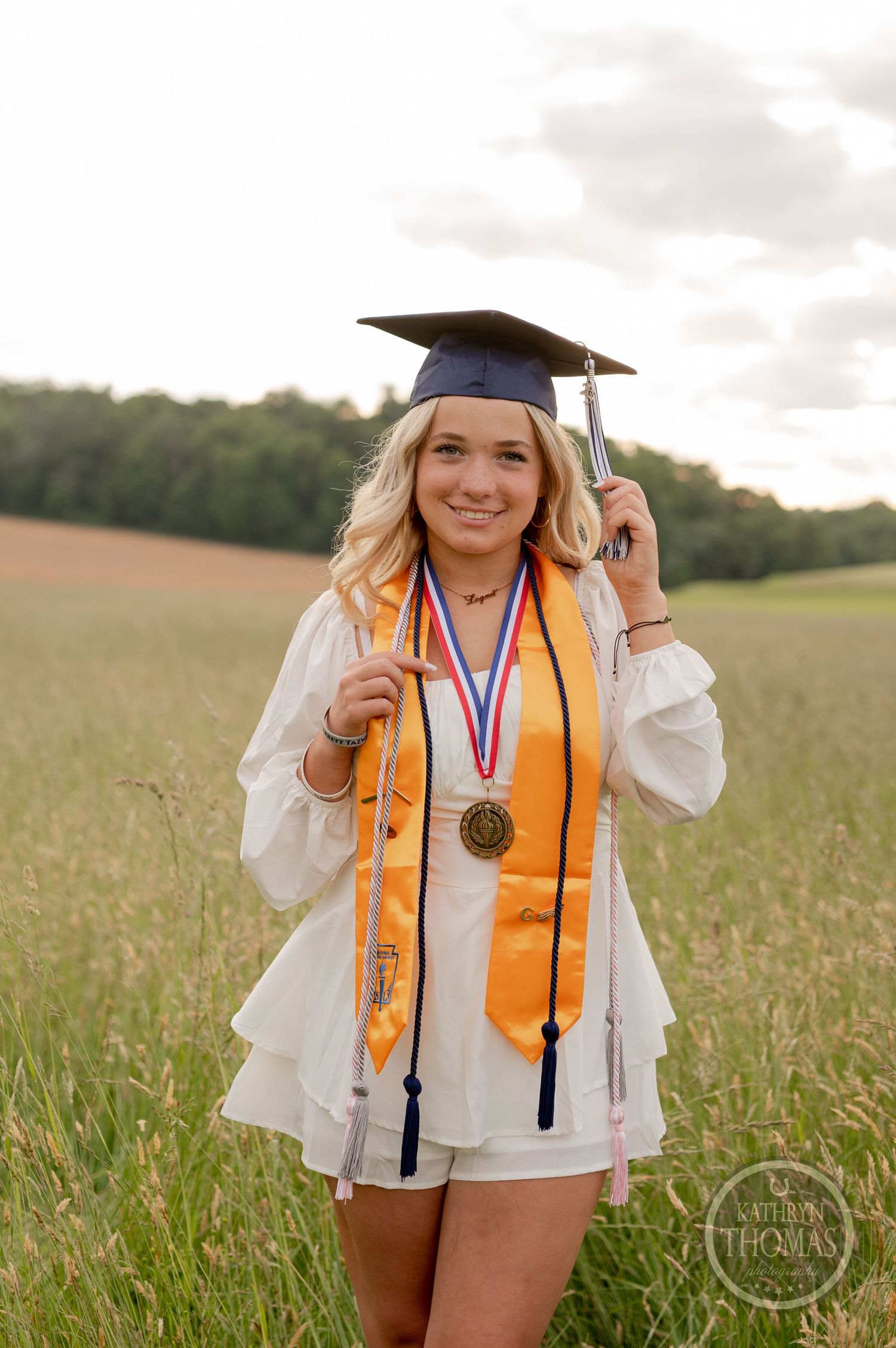 Graduate in white outfit and academic cap with gold stole poses in sunlit meadow.