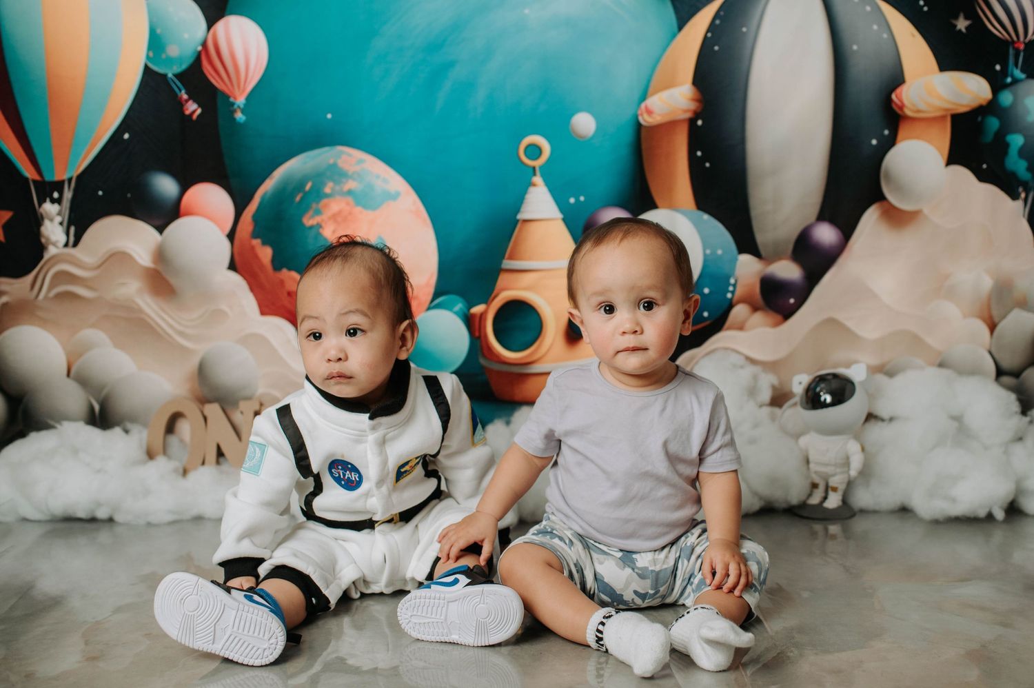 Two babies sit together against a whimsical space-themed backdrop with hot air balloons and planets.