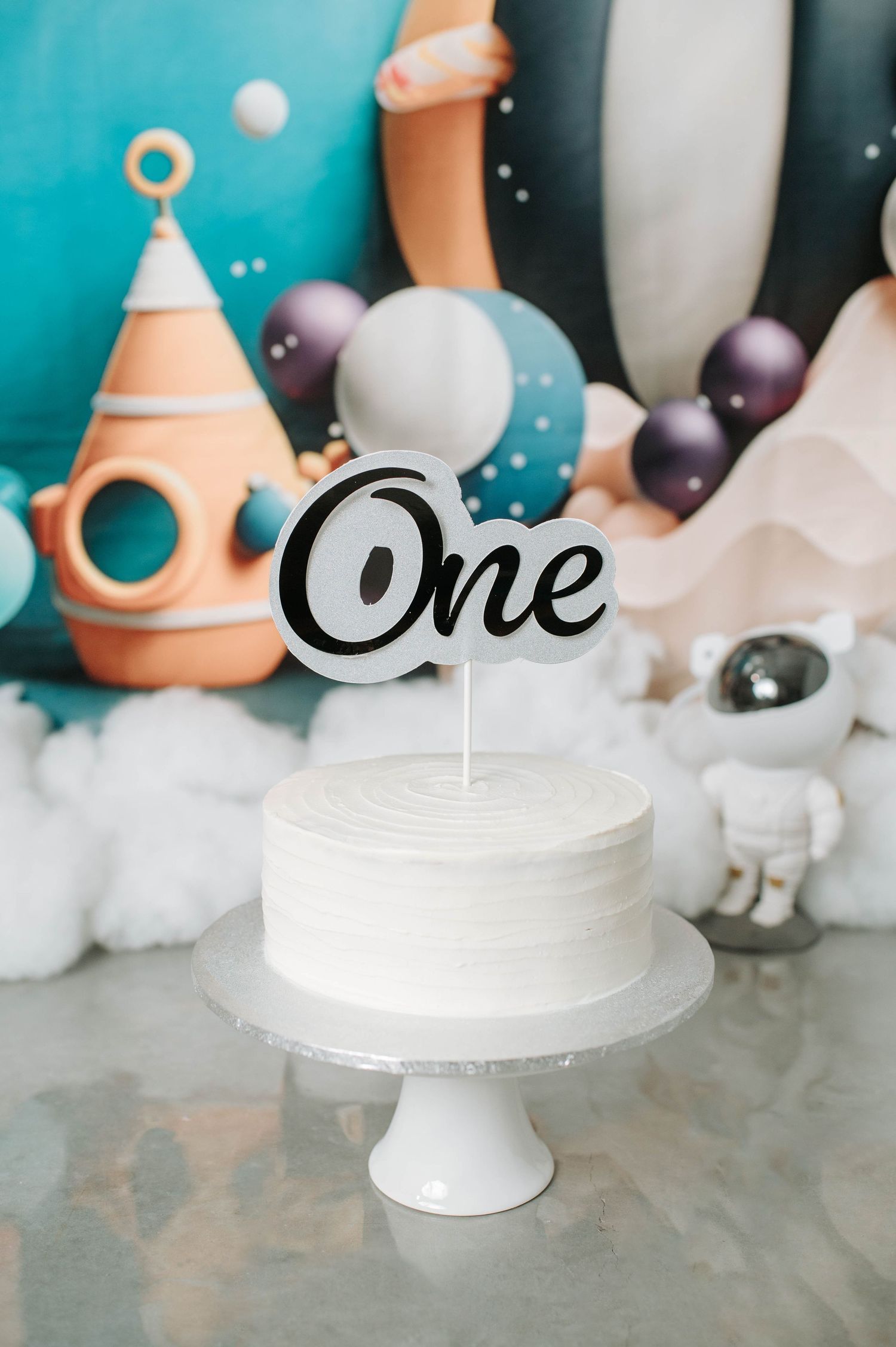 A white birthday cake with 'One' cake topper against a space-themed backdrop with balloons and clouds.