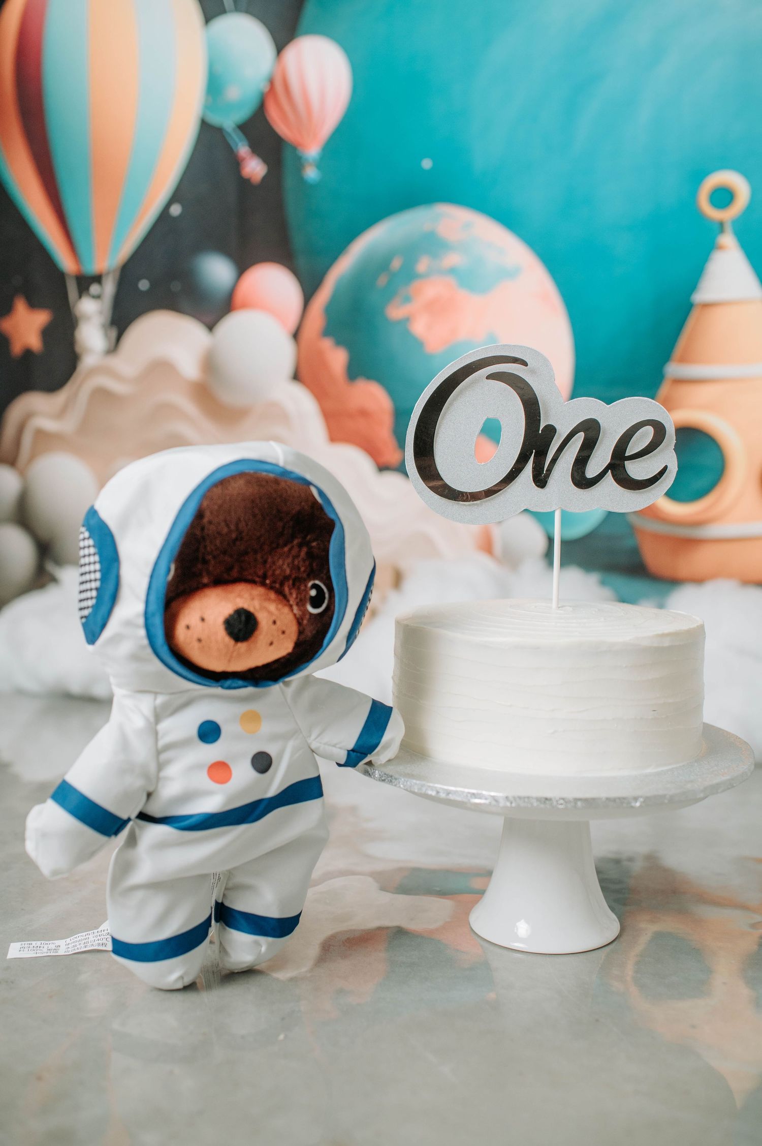 A stuffed teddy bear in an astronaut suit stands next to a white birthday cake with 'One' topper in a space-themed setting.