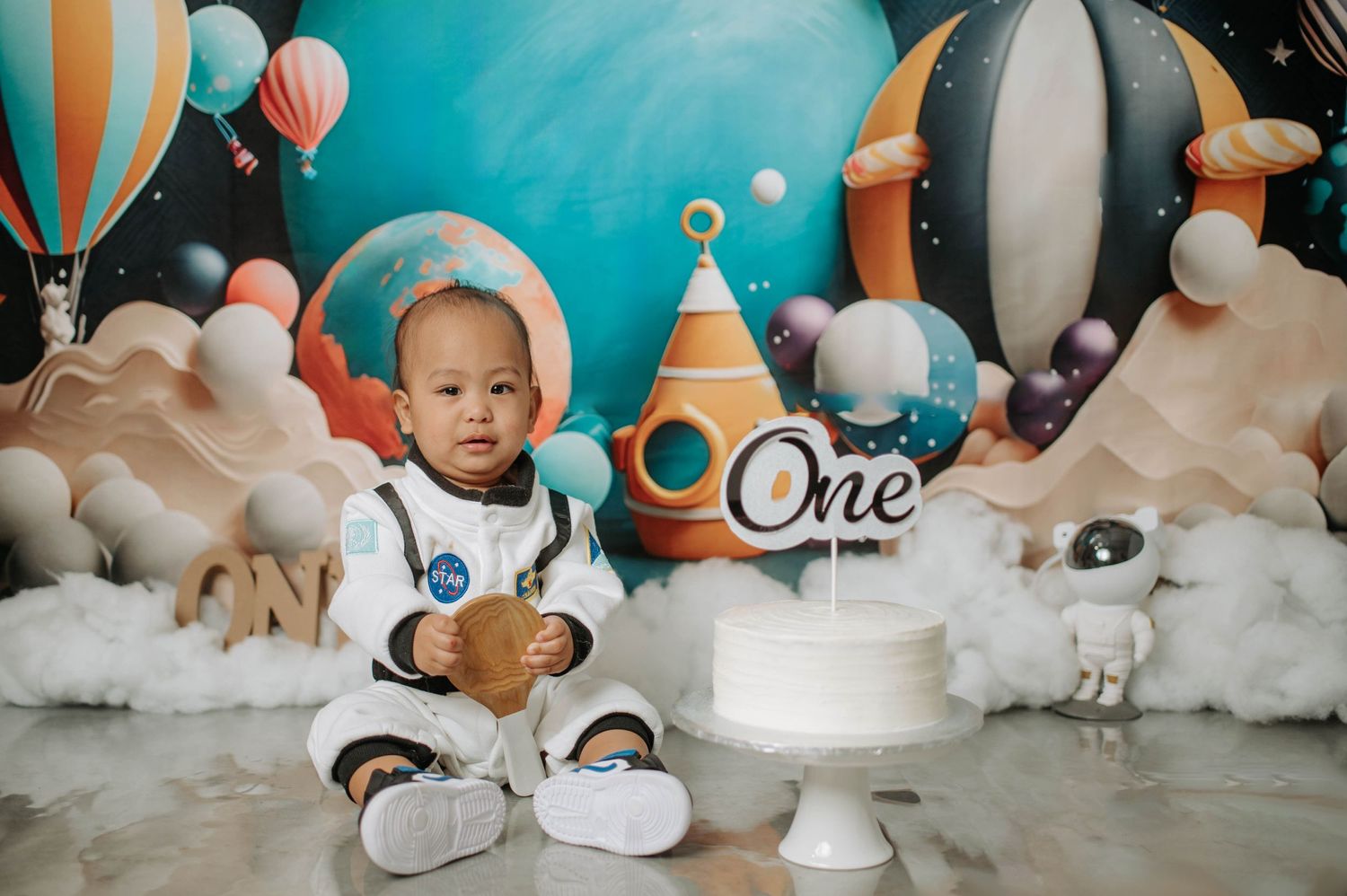 Space-themed first birthday setup with white cake, astronaut decorations, and hot air balloon backdrop.