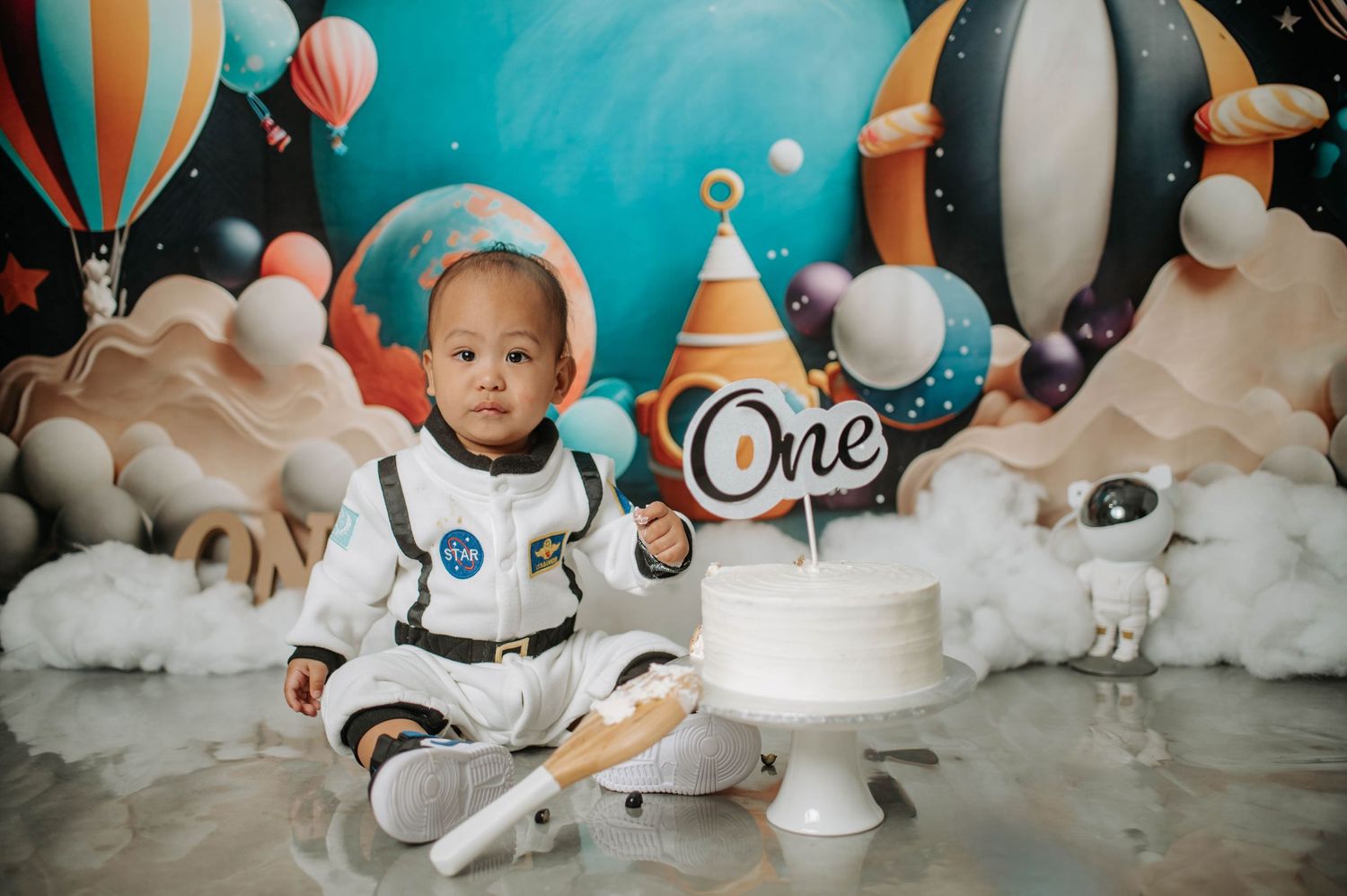 Space-themed first birthday photoshoot with white cake, rocket decorations, and cloudy background.
