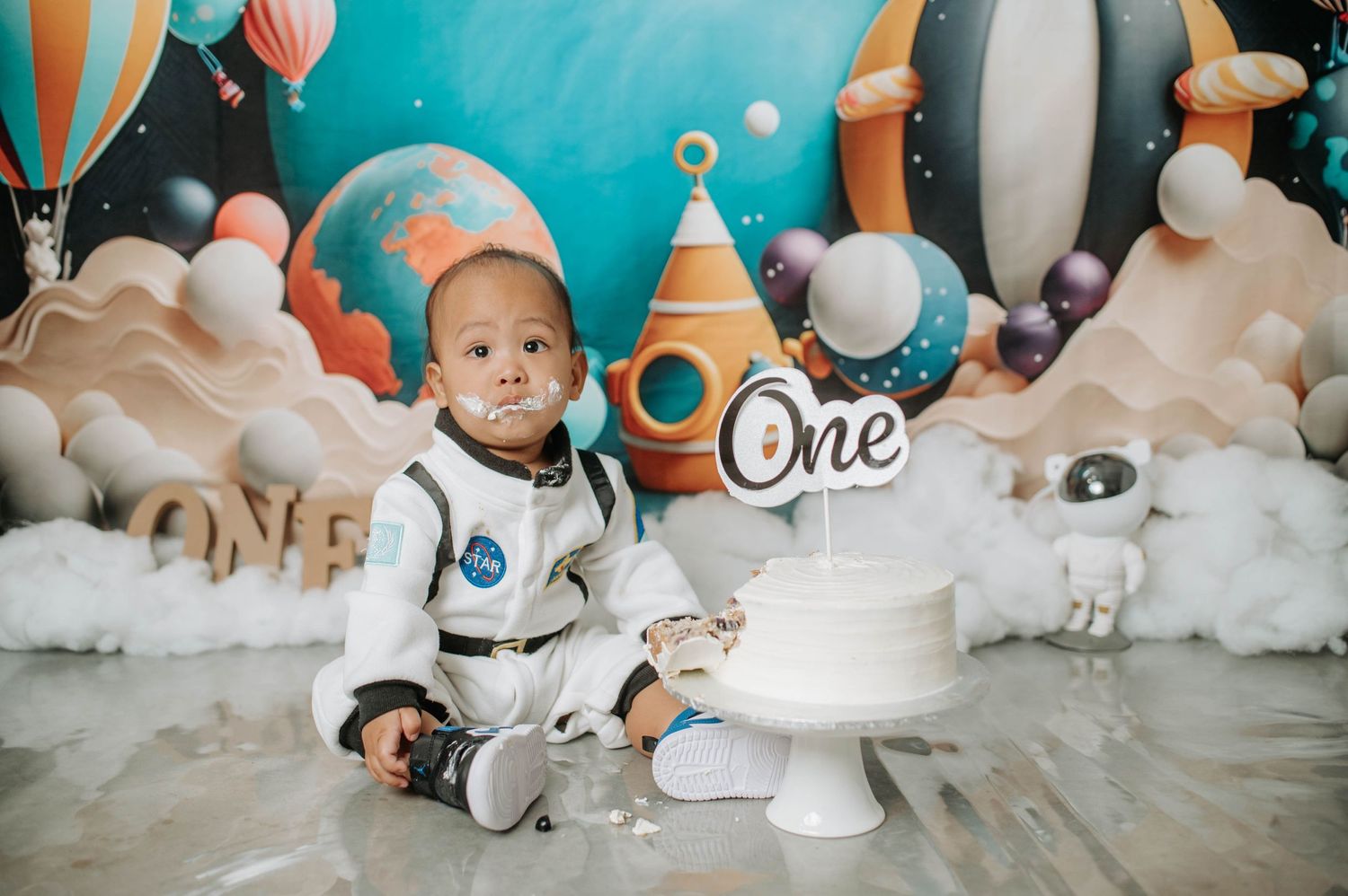 Space-themed cake smash photoshoot with white decorative clouds and hot air balloon backdrop.