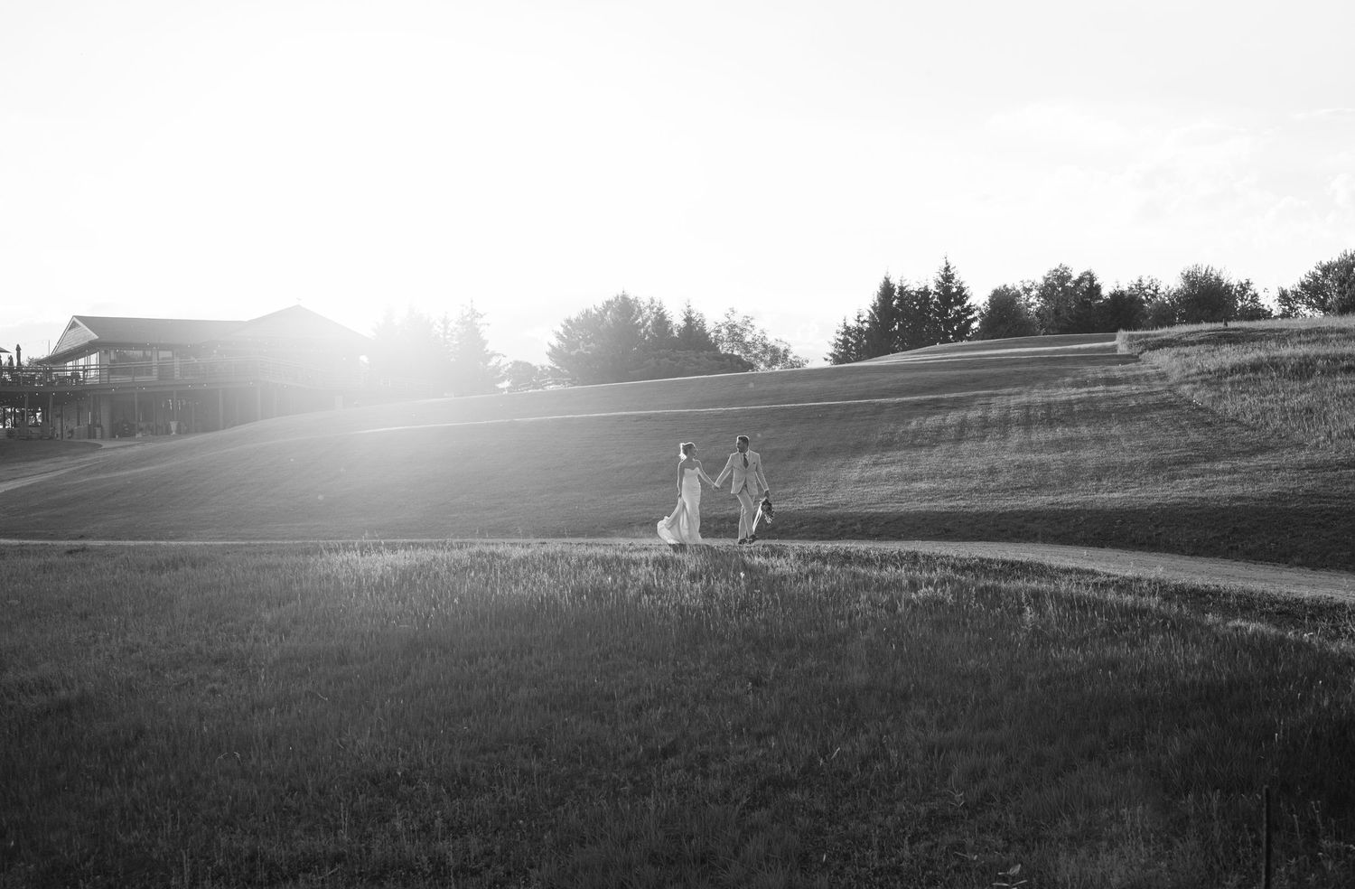 Black and white photo of a couple holding hands walking through a sunlit grassy field with trees and a building in the background.