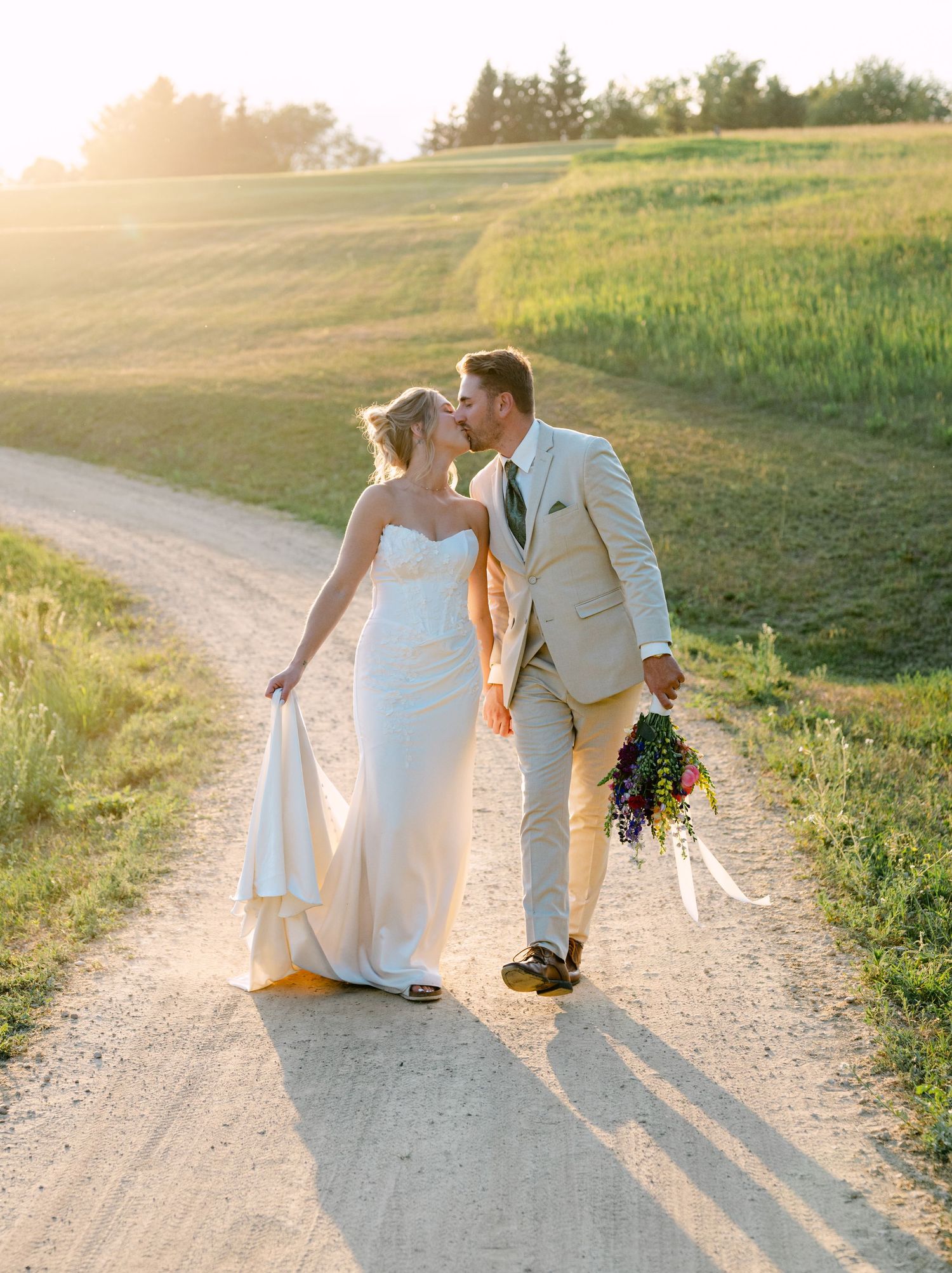 Bride and groom share a golden hour kiss on a scenic dirt road, bride holding colorful bouquet in lush green fields.