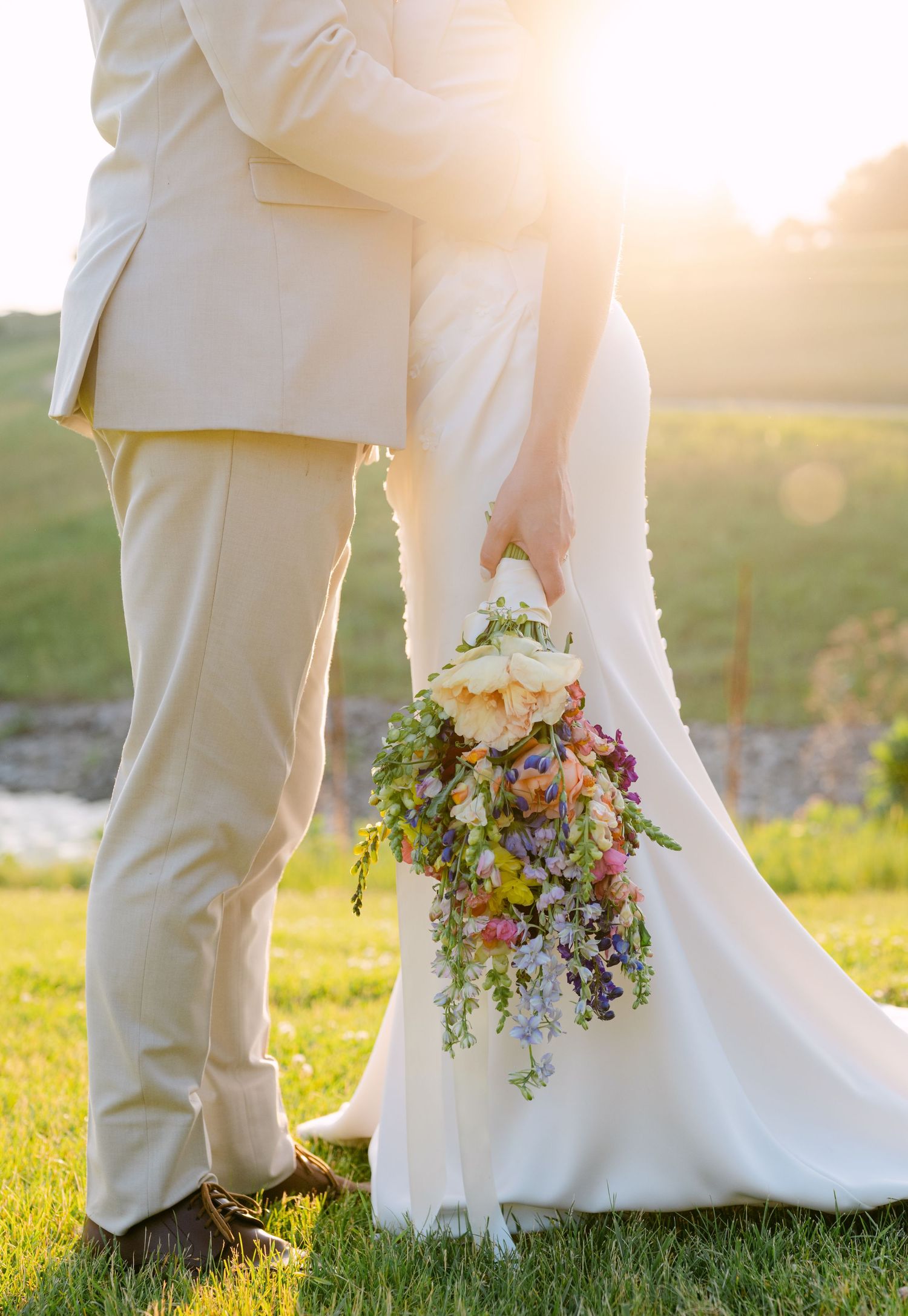 Bride and groom standing on green grass holding a colorful cascading wedding bouquet with golden sunset light.