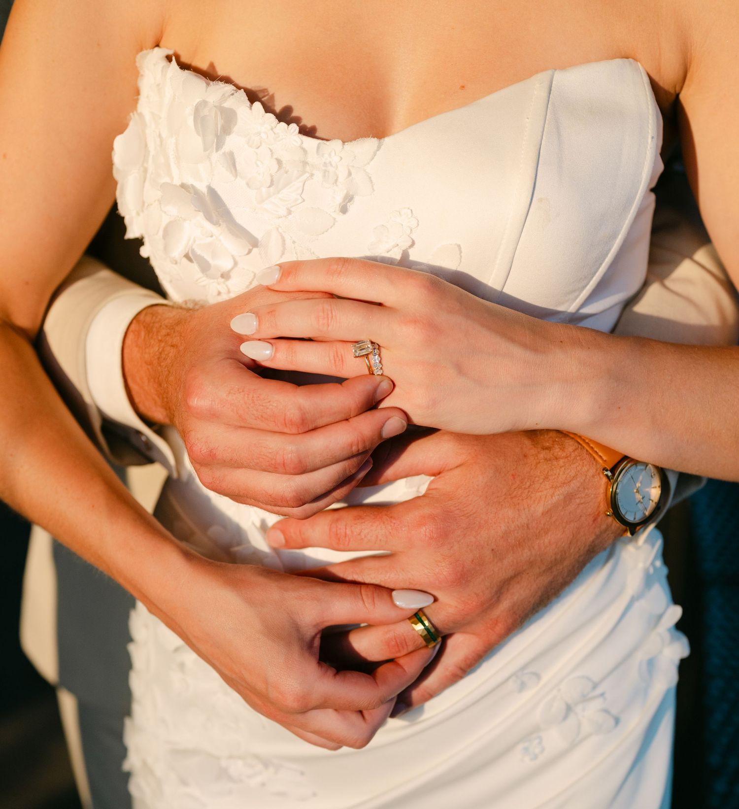 Close-up of newlyweds hands showing wedding rings, bride in white dress with groom wearing a gold watch.