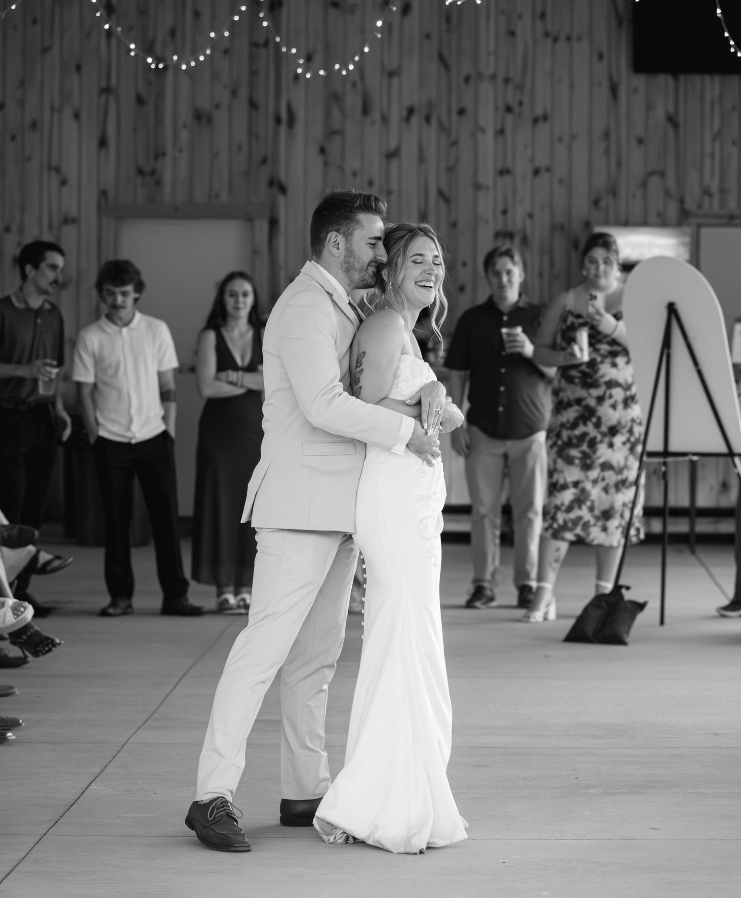 Bride and groom share their first dance in a rustic barn venue while guests watch in black and white photo.
