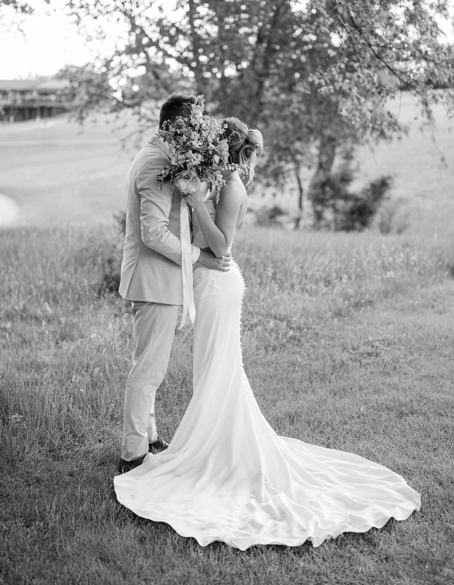 Black and white photo of bride and groom kissing behind a floral bouquet on a grassy lawn.