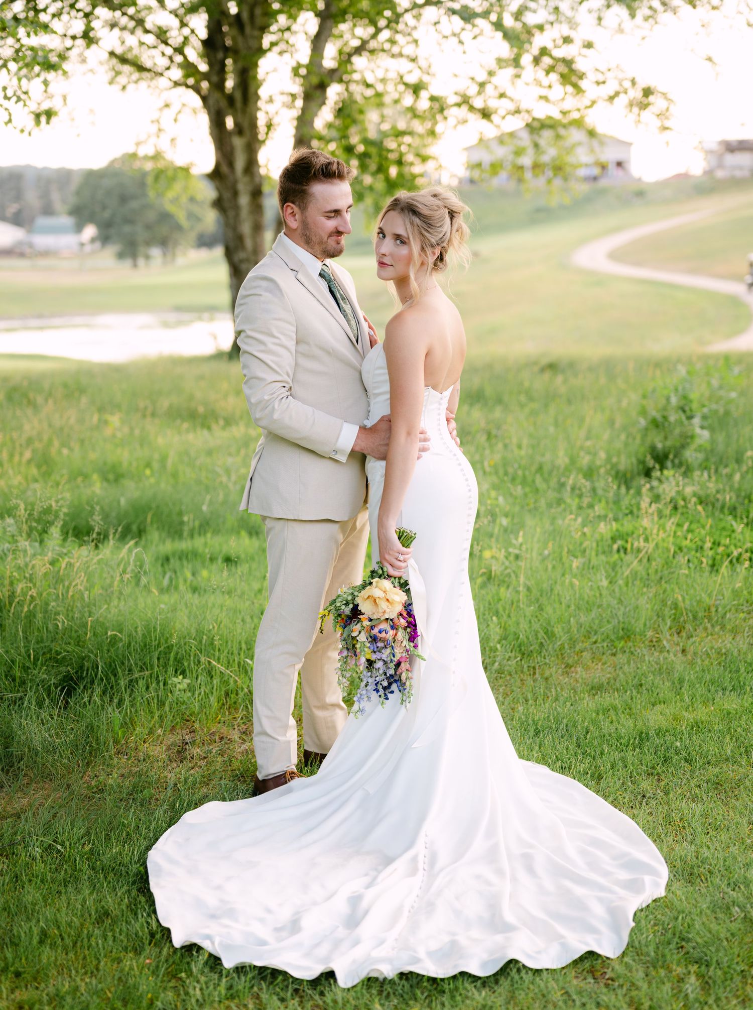Elegant bride in strapless gown with long train poses with groom in tan suit on a green hillside at sunset.